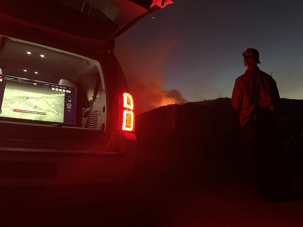 Responder at night beside a vehicle screen showing a live drone map